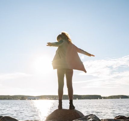 A woman stands on rocks, arms outstretched, enjoying a sunny day by a serene lake.