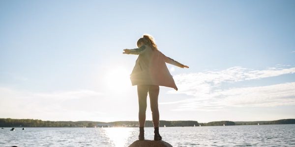 A woman stands on rocks, arms outstretched, enjoying a sunny day by a serene lake.