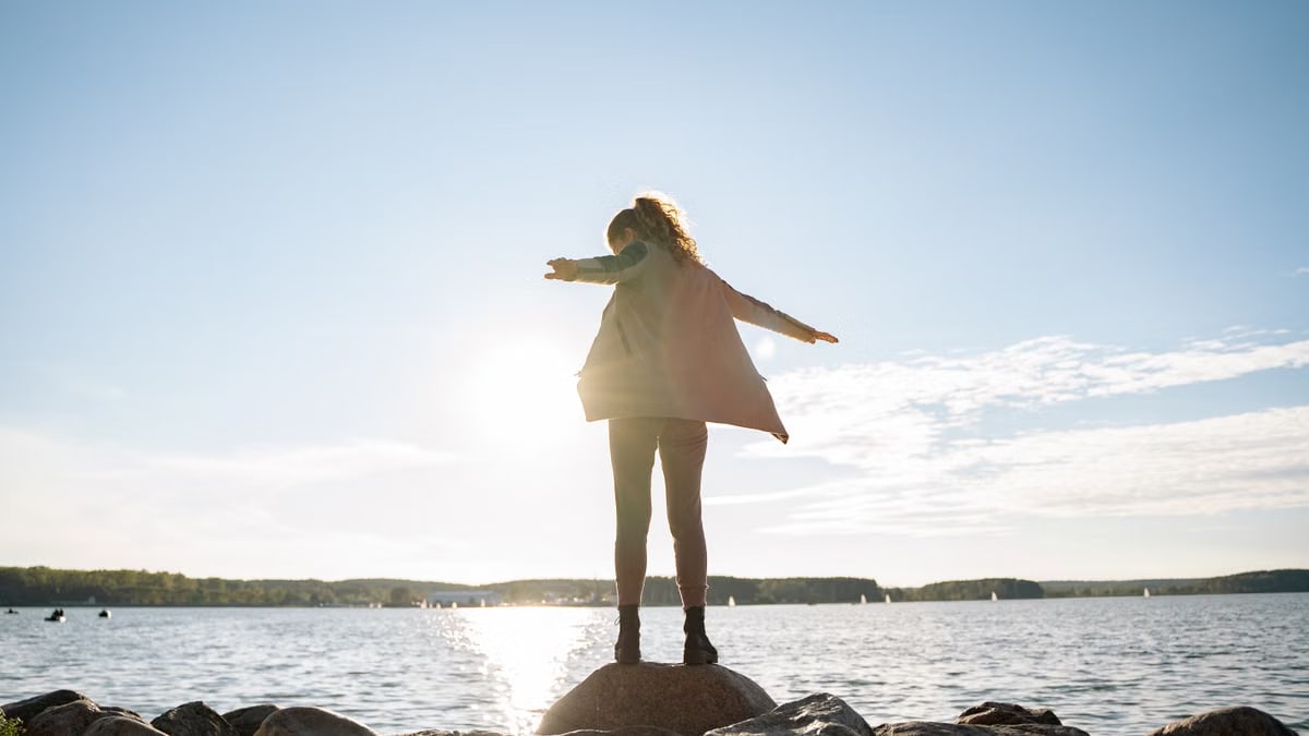 A woman stands on rocks, arms outstretched, enjoying a sunny day by a serene lake.